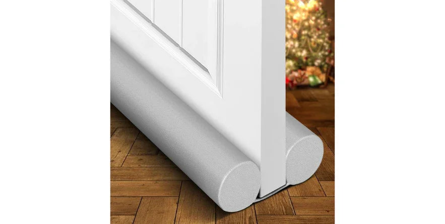 Adjustable gray door draft stopper placed under white door on wooden floor with Christmas tree in background